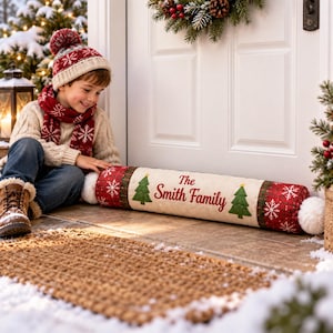 May include: A cylindrical fabric door draft stopper featuring a cream-colored center panel with the red text The Smith Family flanked by two green pine trees. The ends are red with white snowflake patterns and finished with white pom-poms.