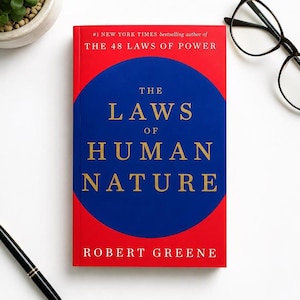 May include: A hardcover book titled The Laws of Human Nature by Robert Greene. The cover features a bright red background with a large blue circle in the center. The title is printed in gold lettering, and the author's name appears at the bottom.