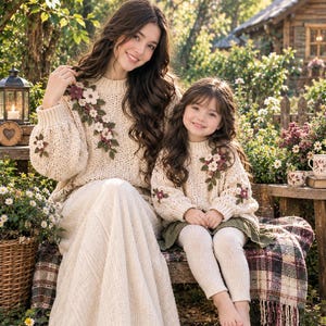 May include: Two matching cream-colored knit sweaters featuring long sleeves and ribbed cuffs. Each sweater is decorated with a cluster of three-dimensional crocheted flowers in shades of pink, white, and green, positioned on the shoulder and sleeve.