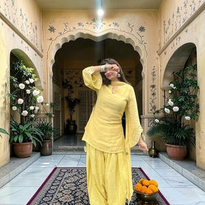 Op de afbeelding: Een felgele salwar kameez-set bestaande uit een tuniek met lange mouwen en uitlopende manchetten en een bijpassende, losvallende broek. Het kledingstuk is voorzien van subtiele borduursels op de mouwen en broek, inclusief een gele dupatta-sjaal.