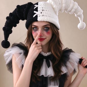 May include: A two-toned jester hat crocheted with black and white yarn, featuring two long, curved, textured horns with pom-poms at the ends and a white ribbon lace-up detail on the front center seam of the cap.