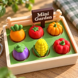 May include: A miniature garden toy set featuring a light beige fence with a brown sign that reads Mini Garden. The base is green with six circular cutouts holding small plastic vegetables: an orange carrot, red tomato, orange pumpkin, purple eggplant, yellow corn, and red bell pepper.