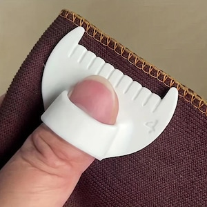 May include: A white plastic sewing tool with a ring for the thumb and a curved, serrated edge designed for guiding fabric. The tool is positioned against a piece of dark brown fabric that features a finished orange stitched edge.