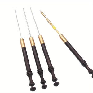 May include: Four needle tools with dark brown wooden handles, brass ferrules, and thin metal shafts. One tool features a series of small, multi-colored beads threaded onto the metal shaft, while the other three tools have plain, bare metal shafts.