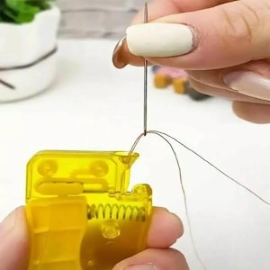 May include: A bright yellow plastic needle threader tool with a small internal metal spring mechanism. The device is shown in use, with a thin silver sewing needle positioned above the funnel-shaped opening, pulling a fine thread through the eye.