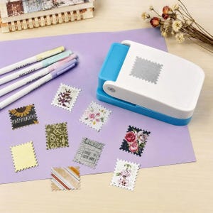 May include: A white and blue paper punch tool with a rectangular base, positioned next to three pastel-colored pens and several small, rectangular paper cutouts with scalloped edges featuring various floral, striped, and patterned designs.