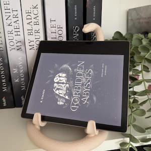 Könnte beinhalten: Ein schwarzer E-Reader zeigt das Buchcover Forbidden Abysses von R. Kimara mit einer Gehirn- und Schlangenillustration. Das Gerät steht auf einem hellbeigen, kreisförmigen, strukturierten Ständer mit einem Schlangenkopf oben in der Mitte.