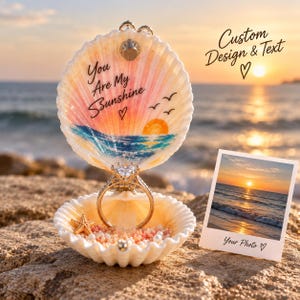 May include: A hinged seashell ring box featuring a painted sunset, ocean waves, and the text "You Are My Sunshine" with a heart. Inside, a silver ring with a clear stone sits among small beads and a tiny starfish. A photo print shows a sunset.