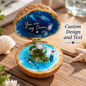 May include: A walnut shell ring box featuring a blue resin ocean interior with a miniature island, a lighthouse, and a whale. The lid interior displays the text You are my Ocean with a small heart and a star. A silver ring sits on the island.