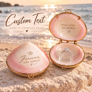 May include: A pink and white seashell ring box with gold trim. The closed shell features the text "Forever and Always" with heart icons. The open shell displays a silver diamond ring and the text "Will You Be Mine?" with a heart icon.