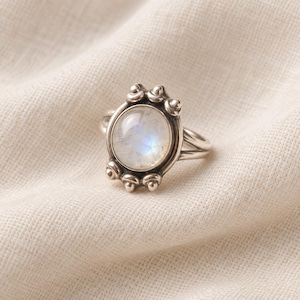 Op de afbeelding: Een zilveren ring met een ronde, doorschijnende maansteen cabochon gezet in een gepolijste metalen zetting. De zetting is versierd met kleine, afgeronde zilveren bolletjes aan de boven- en onderkant, bevestigd aan een eenvoudige, gladde, gespleten ring.