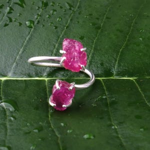 Peut inclure: Une bague ouverte en argent ornée de deux pierres brutes et non polies d'un rose vif, maintenues par des sertis à quatre griffes. L'anneau en métal est lisse et brillant, contrastant avec les surfaces texturées et irrégulières des pierres.