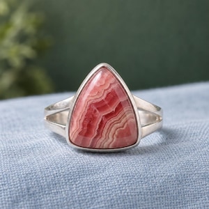 May include: A silver ring featuring a triangular-shaped rhodochrosite gemstone. The stone displays a distinct pattern of alternating pink and white bands. The metal band is smooth and polished, with a split shank design that meets at the stone setting.