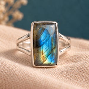 Puede incluir: Un anillo de plata con una gran piedra preciosa de labradorita rectangular de superficie pulida. La piedra muestra destellos iridiscentes en azul y dorado. La banda de metal tiene un diseño de doble brazo que se une al engaste de la piedra.