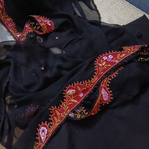 May include: A black sheer fabric scarf featuring a wide, scalloped border in deep red. The border is decorated with intricate floral embroidery in shades of pink, purple, and gold, finished with a delicate black lace trim along the edges.