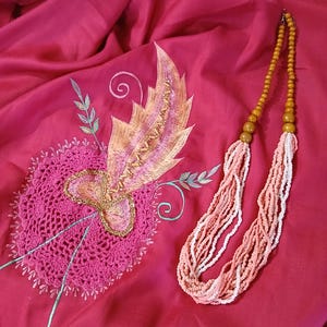 Könnte beinhalten: Ein magentafarbener Stoff mit einer kreisförmigen, rosa Häkelapplikation und gestickten Blattmotiven in Gold, Orange und Grün. Darauf liegt eine mehrsträngige Halskette aus kleinen pfirsichfarbenen und weißen Perlen sowie größeren senfgelben Holzperlen.