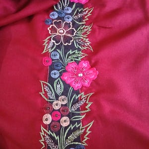Könnte beinhalten: Ein tiefroter Stoff mit einem vertikalen schwarzen Einsatz, der mit aufwendigen Blumenstickereien verziert ist. Das Design umfasst leuchtend rosa Blüten, grüne Blätter und Gruppen kleiner kreisförmiger Elemente in Blau-, Rot- und Pfirsichtönen mit Perlen.
