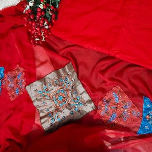 May include: A vibrant red fabric featuring several embroidered patches in square and triangular shapes. The patches display intricate floral and geometric patterns in shades of blue, orange, and white. A small cluster of red berries is visible.