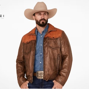 May include: A brown leather jacket featuring a contrasting tan leather yoke across the shoulders, a front zipper closure, and two chest pockets. The jacket is worn over a blue button-down shirt and dark blue jeans with a large metallic belt buckle.