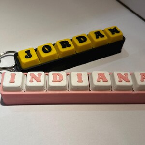 May include: Two rectangular keychains featuring mechanical keyboard-style keys. One keychain has a black base with yellow keys spelling JORDAN in black letters. The other has a pink base with white keys spelling INDIANA in pink letters.