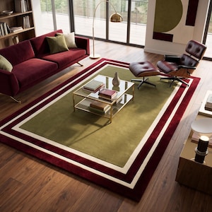 May include: A rectangular area rug featuring a large olive green center panel bordered by a thick white stripe and a wider deep burgundy outer frame. The rug is positioned on a wooden floor beneath a glass coffee table with a gold metal frame.