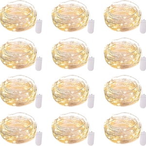 May include: A set of twelve coiled strands of warm white LED fairy lights, each featuring a thin silver-colored wire and a small white rectangular battery pack. The lights are arranged in a grid pattern, showcasing the glowing illumination.