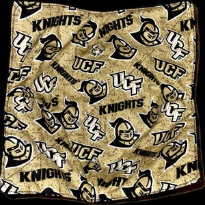 May include: A square, quilted bowl cozy featuring a repeating pattern of the UCF Knights logo and the word "KNIGHTS" in black on a gold background. The cozy has a black border.