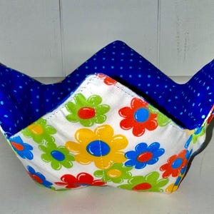 May include: A fabric bowl with a white exterior covered in colorful flowers. The interior is blue with white polka dots. The bowl has a unique shape with pointed edges, perfect for holding food.