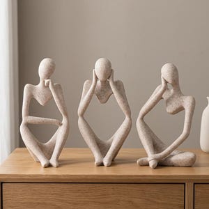 Könnte beinhalten: Drei abstrakte, menschenähnliche Skulpturen mit gesprenkelter beiger Oberfläche. Jede Figur nimmt eine sitzende, nachdenkliche Haltung ein, wobei die Gliedmaßen so angeordnet sind, dass sie offene Zwischenräume bilden. Die Körper sind glatt.