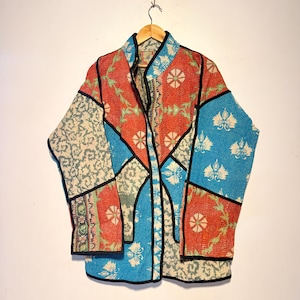 Pode incluir: Um casaco de manga comprida com um design de patchwork de vários padrões florais e geométricos em tons vibrantes de vermelho, azul e creme. A peça é construída com costuras visíveis e acabada com debrum preto ao longo das bordas.