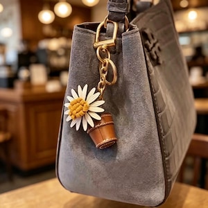 Può includere: Una borsa in camoscio grigio con pannello laterale stampato a coccodrillo presenta un portachiavi in metallo dorato. Il portachiavi include una margherita bianca con un centro giallo senape testurizzato e un piccolo ciondolo in pelle marrone a secchiello.