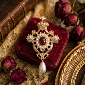Op de afbeelding: Een goudkleurige broche met een centrale ovale donkerrode steen omringd door een ring van kleine witte parels. Het gedetailleerde metalen frame bevat stervormige accenten, bloemmotieven, een kroonvormige bovenkant en een druppelvormige parelhanger.