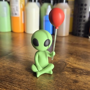 Può includere: Una piccola statuetta aliena verde chiaro seduta a gambe incrociate. L'alieno ha una testa grande, liscia e arrotondata con due grandi occhi neri ovali. Tiene in mano un'asta sottile e trasparente sormontata da un singolo palloncino rosso opaco.