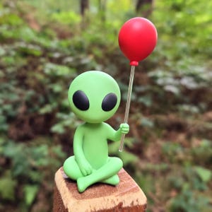 May include: A small green alien figurine with large black oval eyes, sitting in a cross-legged position. The alien holds a thin, clear plastic rod topped with a smooth, round, bright red balloon in its right hand.