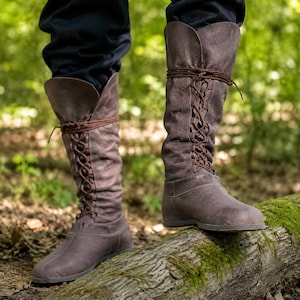 May include: A pair of knee-high brown leather boots featuring a textured surface, a rounded toe, and a front lace-up closure with thin brown laces that wrap around the upper shaft. The boots have a flat sole and a tall, flexible leather construction.