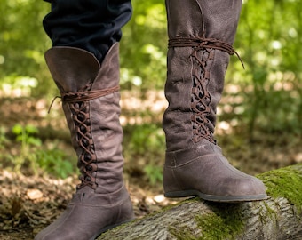 Puede incluir: Un par de botas altas de cuero marrón con superficie texturizada, punta redondeada y cierre frontal de cordones finos que se envuelven alrededor de la caña. Las botas cuentan con una suela plana y una estructura de cuero alta y flexible.