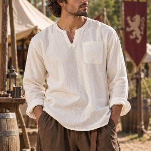 May include: A long-sleeved, off-white shirt featuring a textured fabric, a small chest pocket, and a notched V-neckline. The shirt has a relaxed fit with sleeves that are slightly rolled up at the cuffs, providing a simple and functional design.