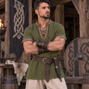 May include: An olive green short-sleeved tunic featuring a V-neckline with brown lace-up detailing and patterned trim on the collar and sleeves. The garment is worn with a wide brown leather belt, a sheath, and dark brown leather wrist cuffs.