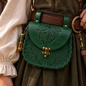 Puede incluir: Un bolso de cuero verde oscuro con un diseño de hoja en relieve en la solapa y patrones de enredaderas en el cuerpo. El bolso tiene un cierre de metal color latón, bordes de cuero trenzado y está sujeto a un cinturón de cuero marrón.