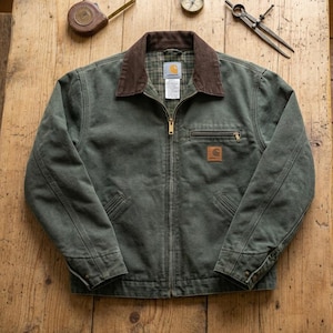 Pode incluir: Um casaco verde-oliva escuro com gola de veludo cotelê castanho e fecho de correr frontal. O casaco apresenta um bolso no peito com fecho, dois bolsos inferiores e um emblema da marca Carhartt. O interior é forrado com tecido xadrez verde.