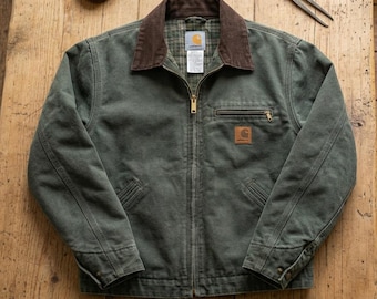 Op de afbeelding: Een donker olijfgroene jas met een bruine corduroy kraag en een ritssluiting aan de voorzijde. De jas heeft een borstzak met rits, twee zakken aan de onderkant en een Carhartt-merklabel. De binnenzijde is gevoerd met een groene ruitjesstof.