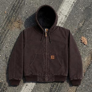Pode incluir: Um casaco com capuz castanho-escuro com fecho de correr frontal, punhos e bainha canelados e um bolso frontal tipo canguru. A peça tem um forro interior preto e uma pequena etiqueta retangular da marca Carhartt no bolso.