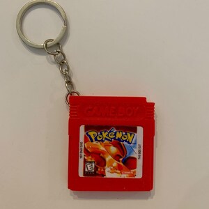 May include: A red Game Boy cartridge keyring featuring a label with the Pokémon logo, an illustration of Charizard, the text DMG-APAE-USA, THIS SIDE OUT, and an E rating symbol. The keyring includes a silver metal ring and a short chain.