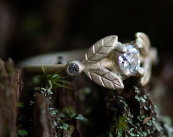May include: A delicate gold ring with a single, clear gemstone surrounded by leaf-shaped details. The ring is resting on a mossy, textured surface.