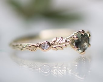 May include: A gold ring featuring a round, dark green gemstone set in a prong setting. The band is textured with a detailed leaf motif and includes a small, round, clear accent stone embedded within the metalwork near the central setting.