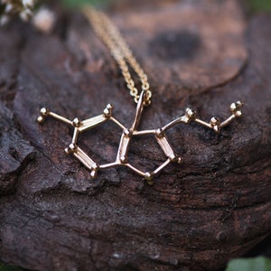 May include: A gold necklace with a pendant shaped like a molecule. The necklace is resting on a piece of wood with green leaves in the background.