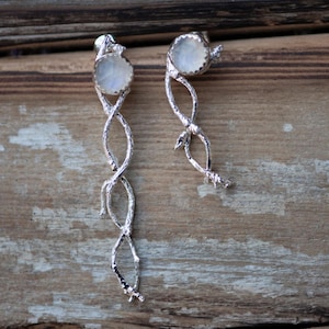 May include: A pair of silver-toned drop earrings. Each earring features a round, iridescent stone at the top, with a twisted, branch-like design extending downwards. The earrings are displayed against a rustic, wooden background.