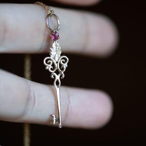 Op de afbeelding: Een goudkleurige sleutelhangerketting met een kleine, ronde, rode edelsteen. Het sleutelontwerp heeft sierlijke krullen en een bladmotief. De ketting wordt door de vingers van een persoon tegen een donkere achtergrond gehouden.