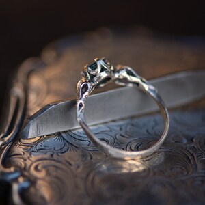 May include: A silver ring with a green gemstone set in a delicate floral design. The ring is resting on a silver tray with an ornate pattern.