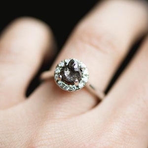 Puede incluir: Un anillo de plata con un gran diamante gris oscuro en bruto rodeado por un halo de diamantes más pequeños y claros.